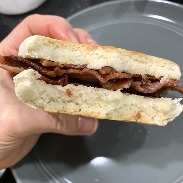 hand holding a gluten free vegan english muffin stuffed with bacon over a grey plate